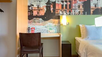 Habitación de hotel con cama, escritorio, silla, un mural en la pared y una mesita de noche.