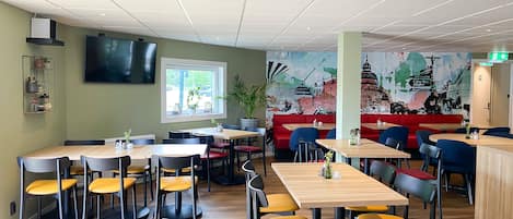Un restaurante moderno con mesas y sillas de madera, un gran mural en la pared y una zona con banquitos rojos.