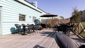 A wooden deck with outdoor furniture, including chairs and a table, adjacent to a light blue building with windows and a chimney. - Good Morning Lund (Lund)
