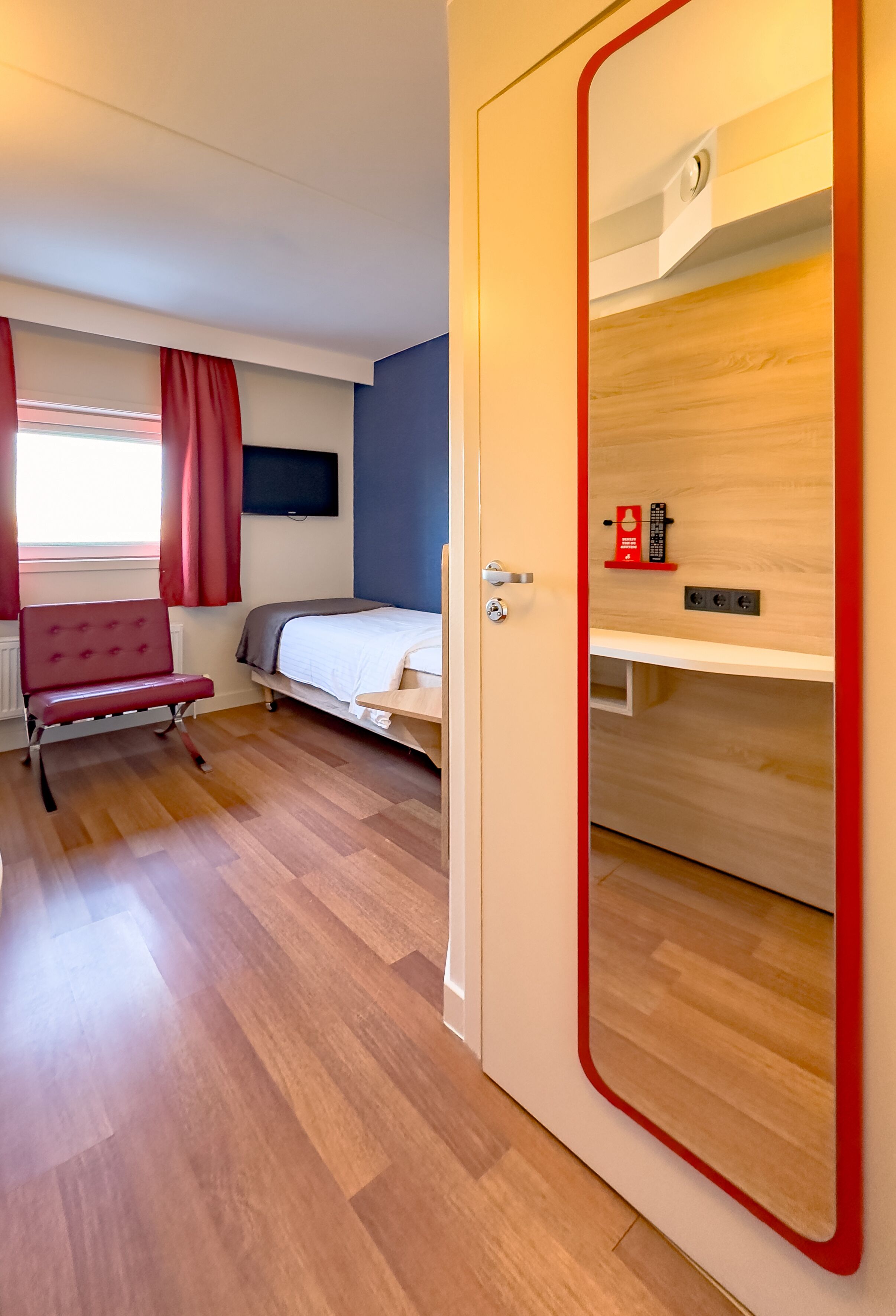 Una habitación de hotel moderna con piso de madera, un sillón tapizado en rojo, una cama, un televisor montado en la pared y un espejo con marco rojo.