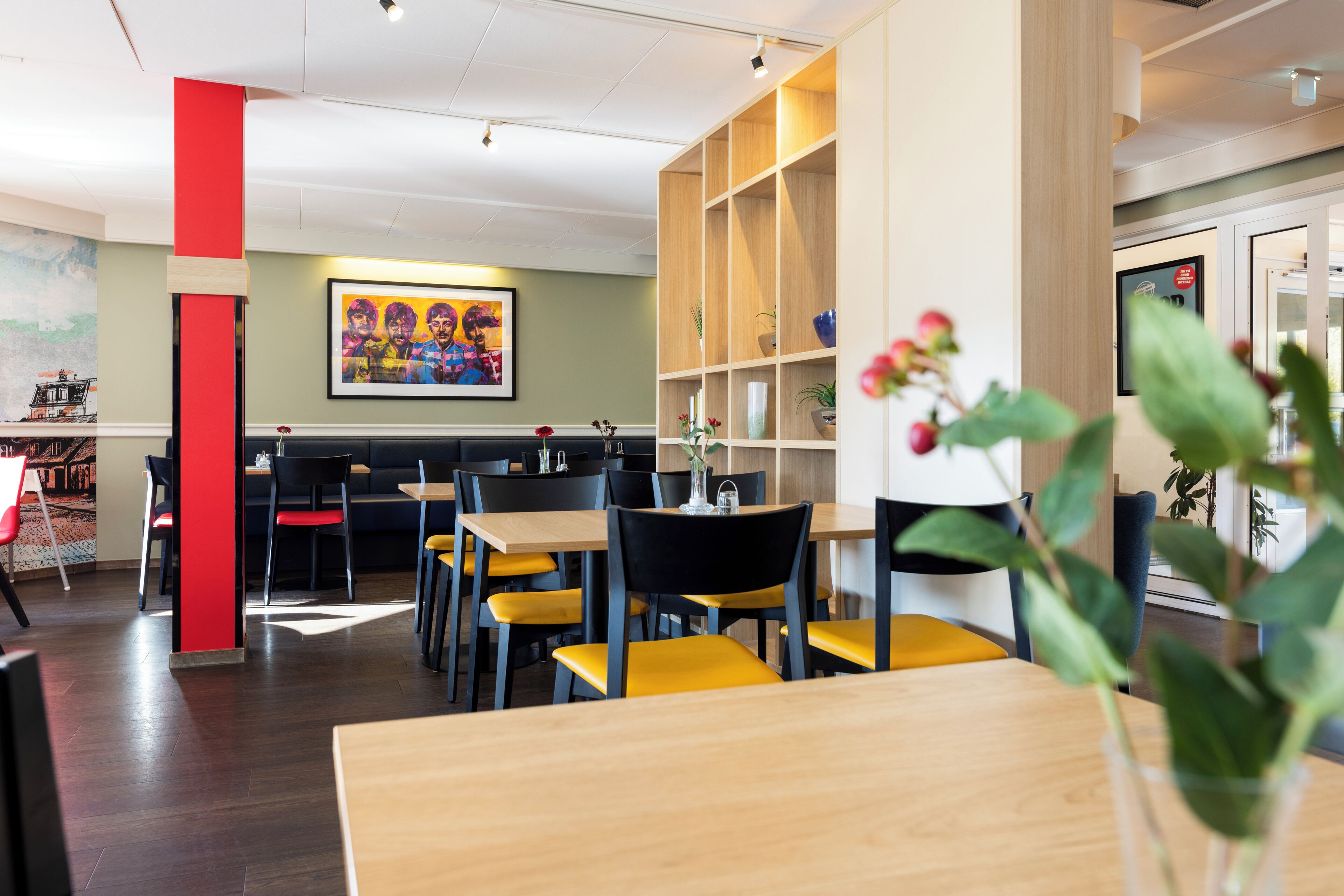 A modern dining area with wooden tables and chairs, a red accent wall, and a colorful painting on the wall.