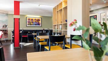 A modern dining area with wooden tables and chairs, a red accent wall, and a colorful painting on the wall.
