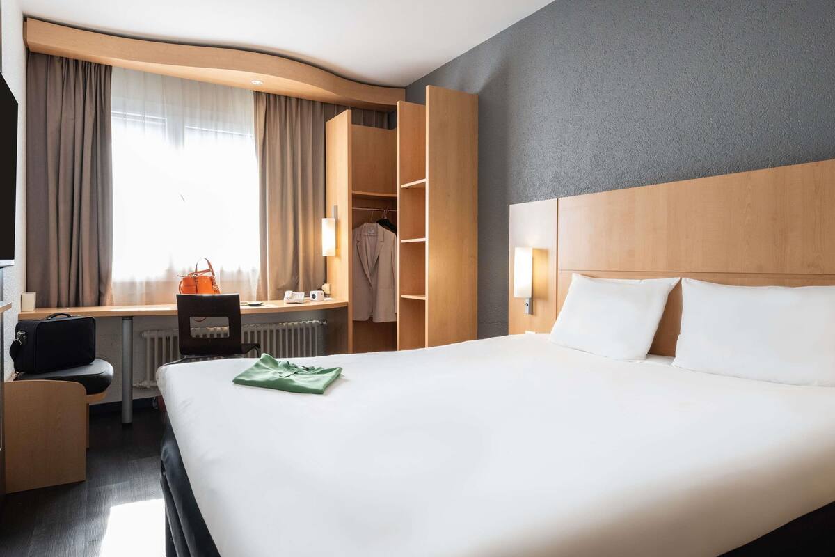 A hotel room with a bed, a desk, a chair, a wardrobe, and a window with curtains.