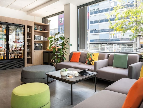 A modern living area with a sofa set, a coffee table, a bar refrigerator, and a large window offering a city view.