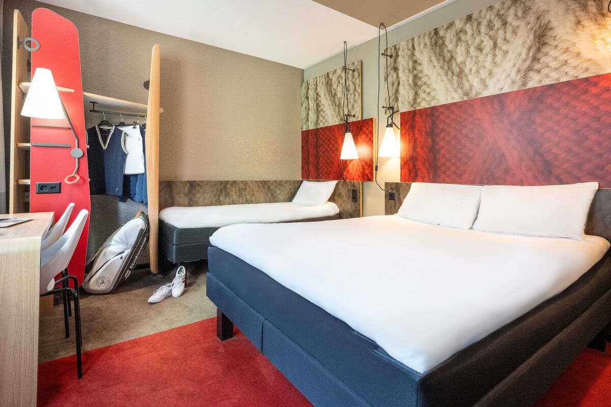 A hotel room with two beds, a desk, a chair, a coat rack, and a red carpet.