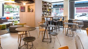 A modern cafe with round tables and wire mesh chairs, a wooden bookshelf, and a view of parked cars outside. - ibis Geneve Centre Gare (Geneva)