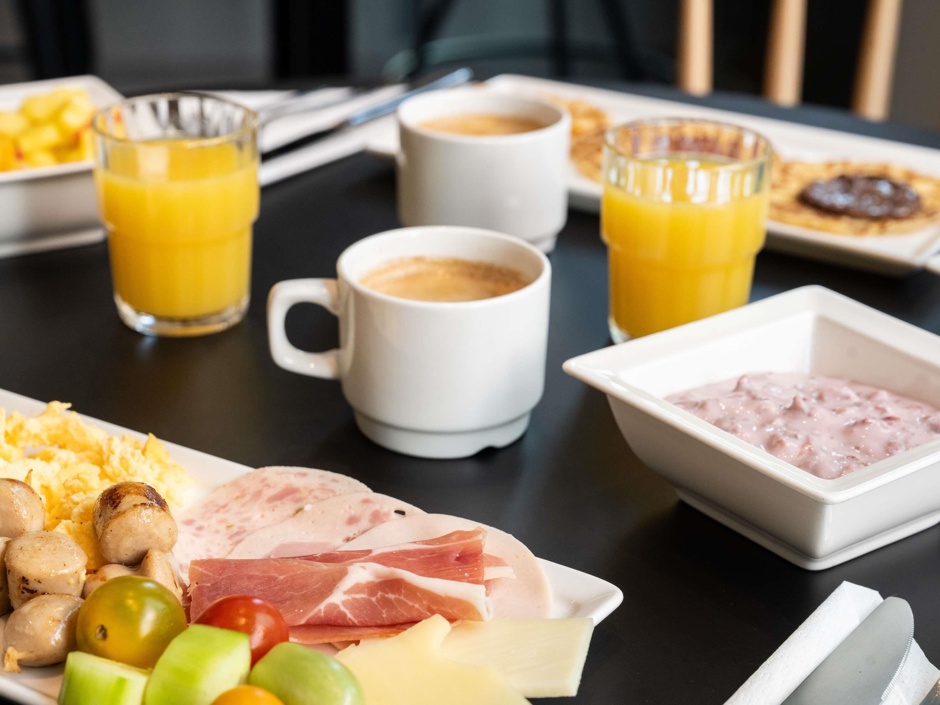 Une table dressée pour le petit-déjeuner avec des œufs brouillés, des saucisses, du jambon, du fromage, des olives et un bol de pâte à tartiner rose. On y trouve également deux verres de jus d’orange et deux tasses de café.