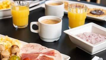 Une table dressée pour le petit-déjeuner avec des œufs brouillés, des saucisses, du jambon, du fromage, des olives et un bol de pâte à tartiner rose. On y trouve également deux verres de jus d’orange et deux tasses de café.