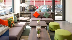 A modern living room with a sofa, ottomans, a coffee table, and a view of parked cars through large windows. - ibis Geneve Centre Gare (Geneva)