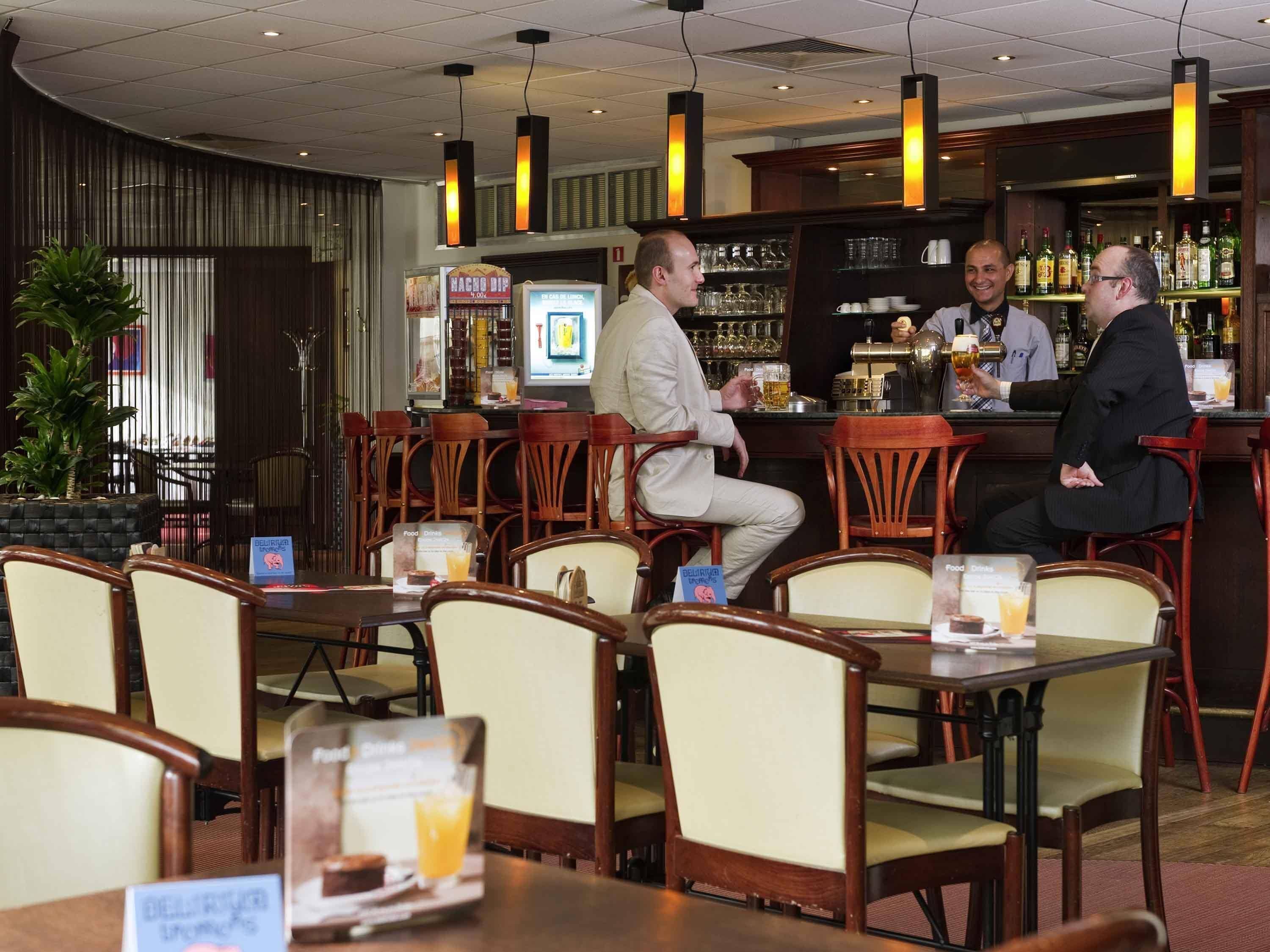 Una zona de bar con sillas y mesas de madera, una barra con un barman y un atril para el menú.