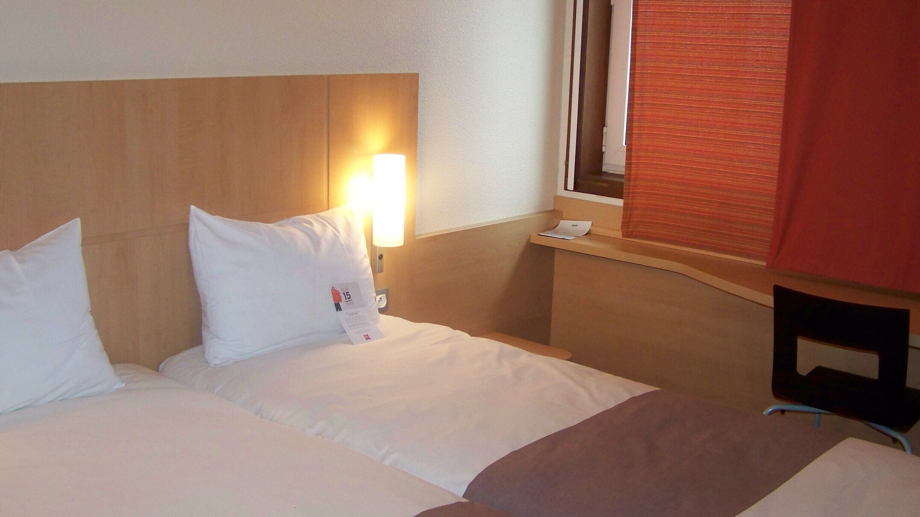Une chambre d’hôtel comprenant un lit, des oreillers, une table de chevet, une lampe, une fenêtre avec des stores et une chaise.