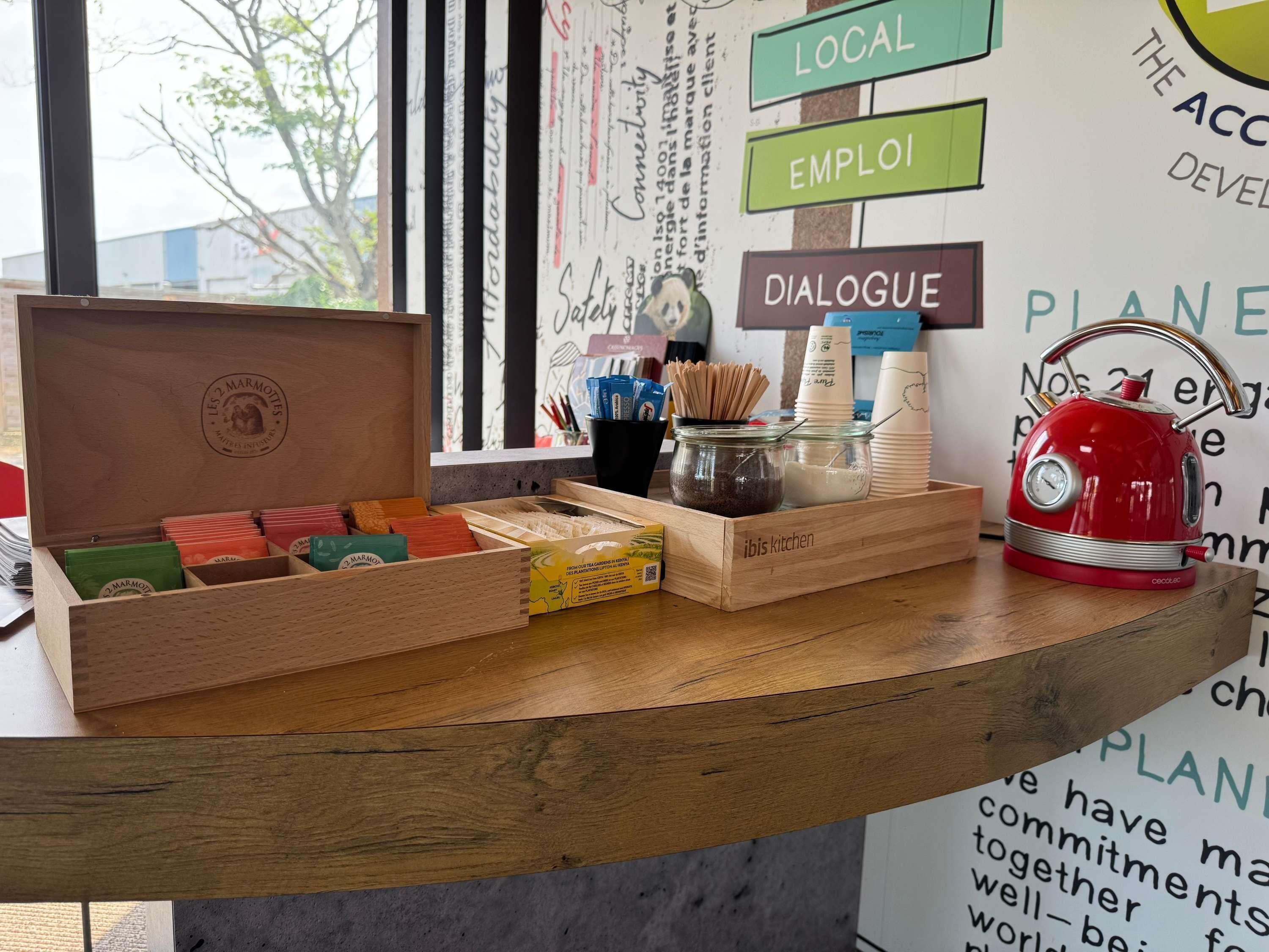 Un comptoir en bois, avec une bouilloire rouge, des bocaux et une boîte de sachets de thé.