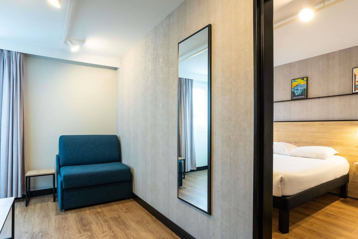 A modern hotel room with a wooden floor, a blue sofa, a bed with white bedding, and a large mirror on the wall.