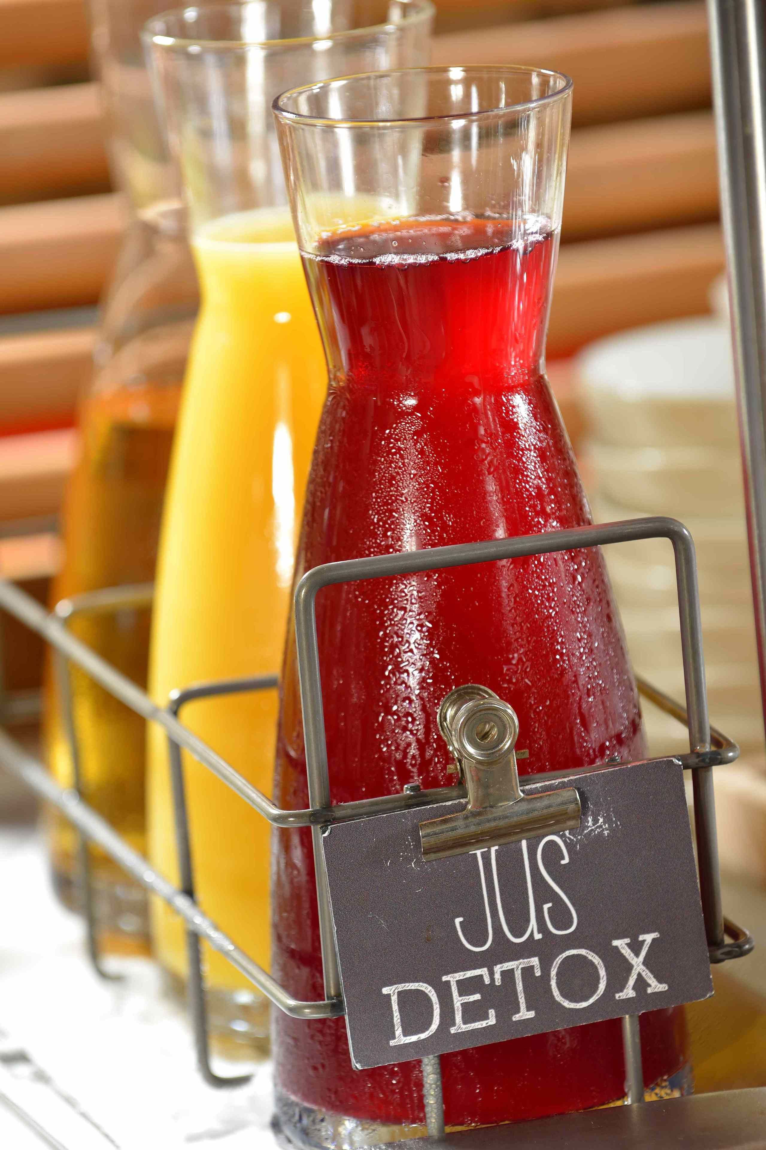 Dispensador de bebidas con una etiqueta que dice "JUS DETOX" y dos vasos de líquidos rojo y naranja.