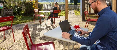 Un área de estar al aire libre techada, con vigas de madera, sillas rojas y una mesa con una computadora portátil y una taza roja.