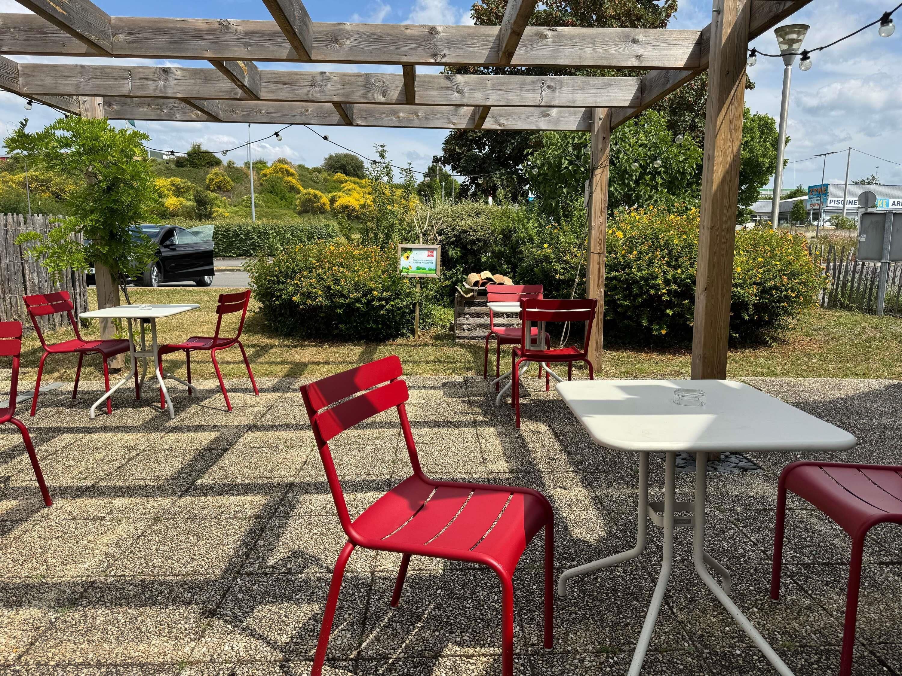 An outdoor patio area with red metal chairs and white tables, surrounded by greenery and a wooden pergola.