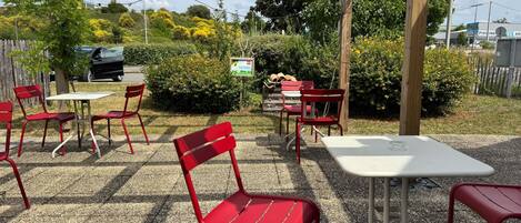 An outdoor patio area with red metal chairs and white tables, surrounded by greenery and a wooden pergola.