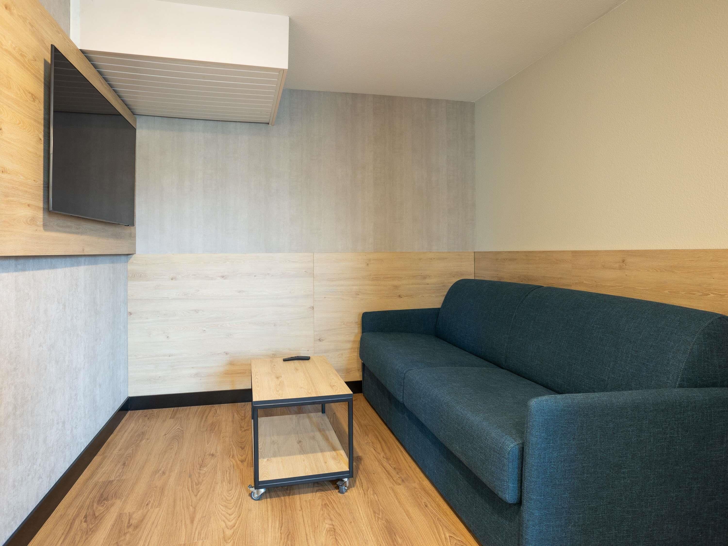 Een kamer met een houten vloer, een blauwe bank, een klein houten tafeltje met een afstandsbediening, en een wandairconditioner.
