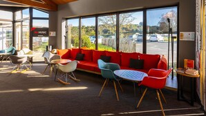 A modern interior with a red sofa, colorful chairs, and large windows offering a view of the parking lot and greenery outside. - ibis Angouleme Nord (Champniers)