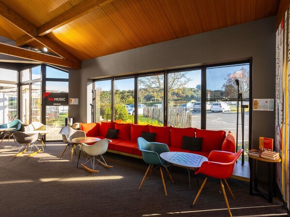 A modern interior with a red sofa, colorful chairs, and large windows offering a view of the parking lot and greenery outside. - ibis Angouleme Nord (Champniers)