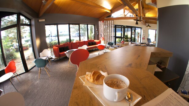 A modern interior with a wooden table, a croissant, and a cup of coffee. There are colorful chairs and a red sofa. - ibis Angouleme Nord (Champniers)