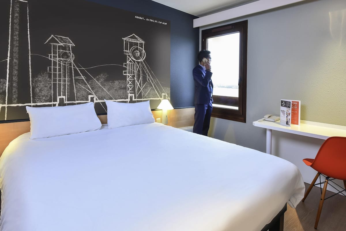 A hotel room with a large bed, a desk with a chair, a window, and a wall with a geometric design.