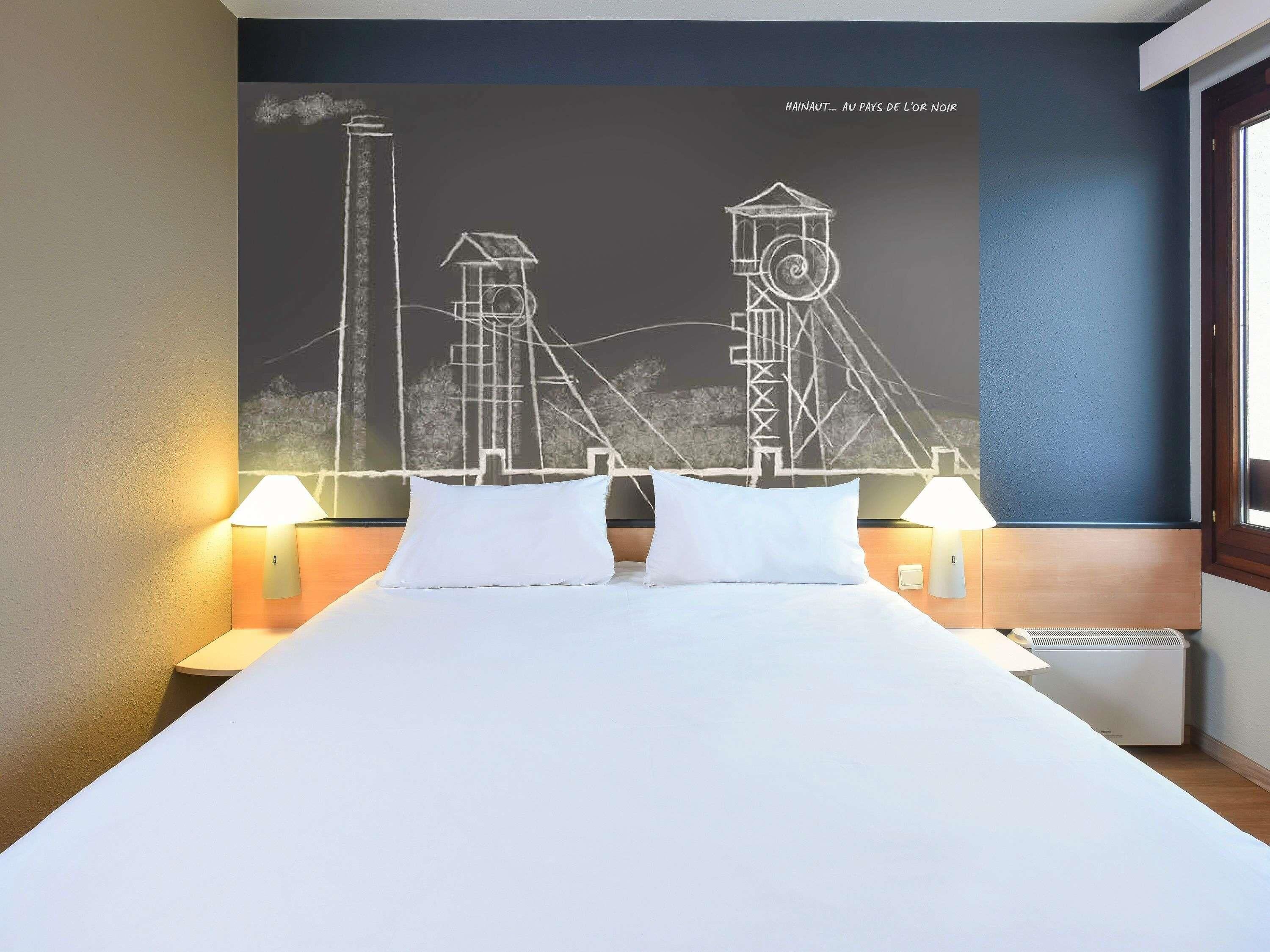 A hotel room with a bed, two bedside lamps, a wall mural, and a window.