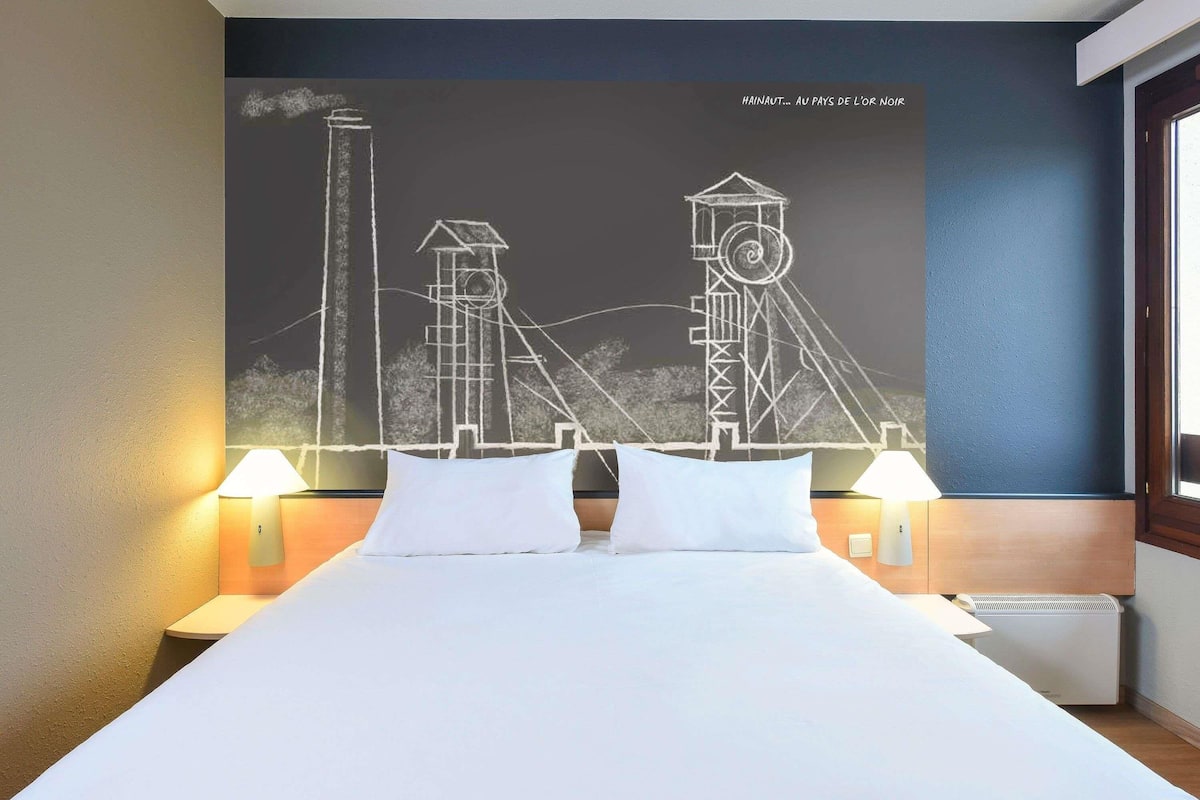 A hotel room with a bed, two bedside lamps, a wall mural, and a window.