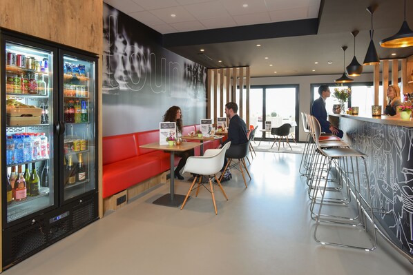 Un café moderne avec un coin salon doté de banquettes rouges, un bar avec des tabourets et un réfrigérateur rempli de boissons.