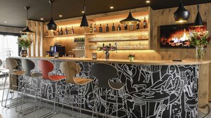 Zona de bar con mostrador de madera, varias lámparas colgantes, un televisor montado en la pared y una variedad de botellas y vasos en estantes.