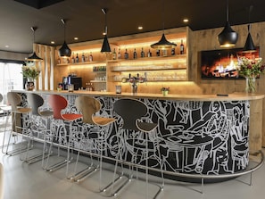 Un espace bar avec un comptoir en bois, plusieurs lampes suspendues, un tĂ©lĂ©viseur fixĂ© au mur, ainsi quâune variĂ©tĂ© de bouteilles et de verres disposĂ©s sur des Ă©tagĂšres.