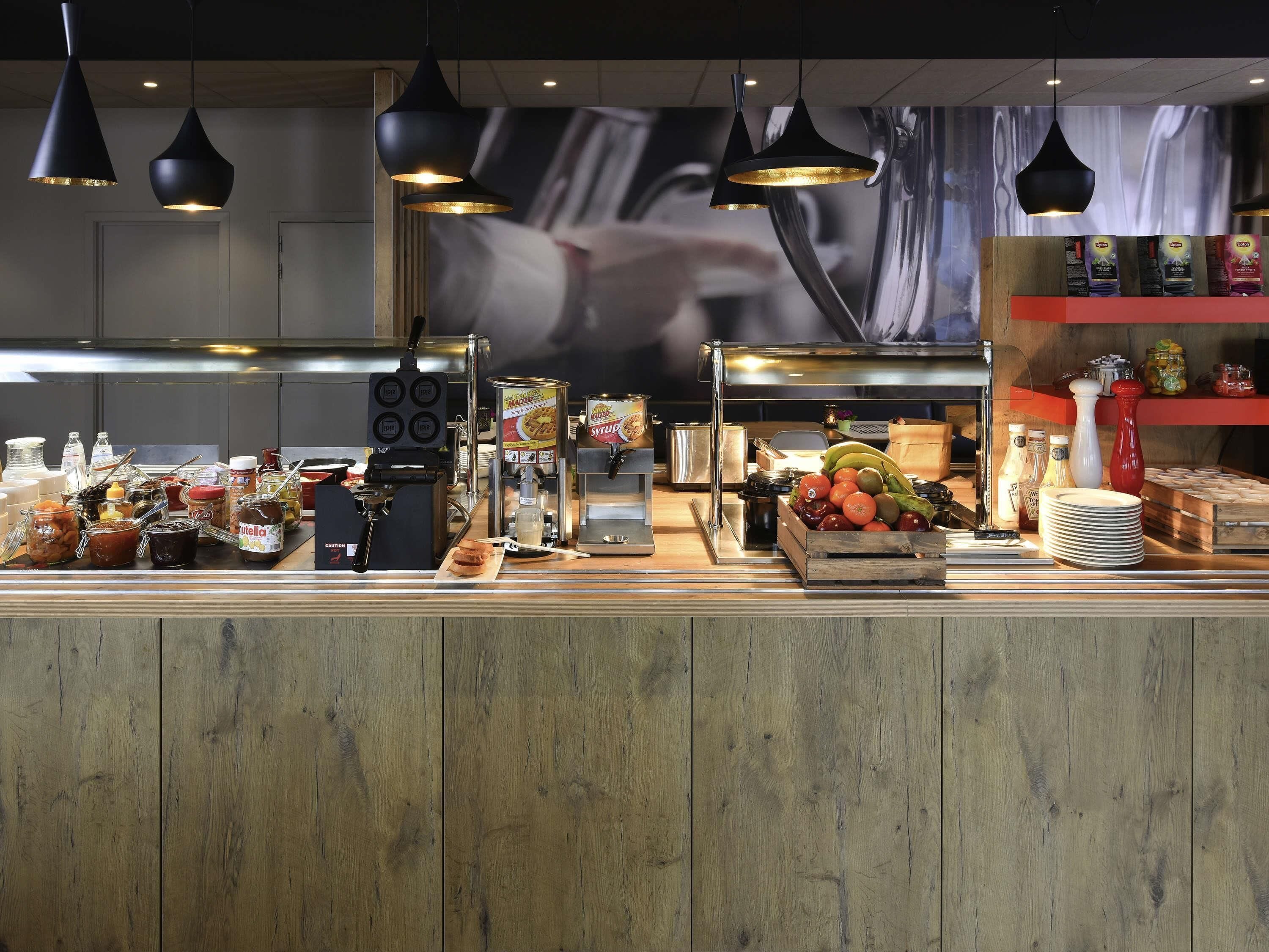 Ein Frühstücksbuffet mit verschiedenen Speisen und Beilagen, einer Kaffeemaschine und hängenden Pendelleuchten.