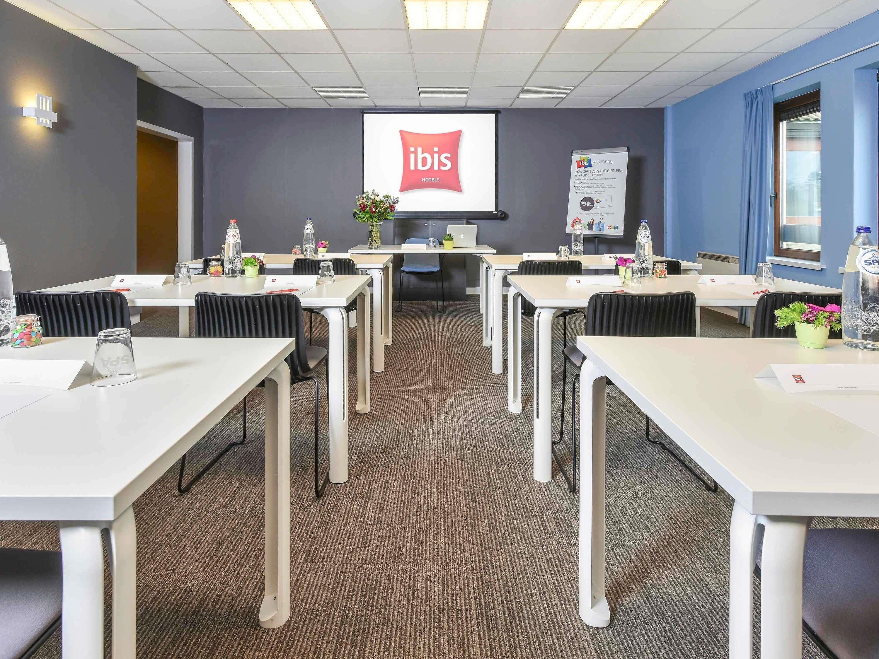 Une salle de conférence avec des tables blanches, des chaises noires, un écran de projection affichant le logo « ibis Hotels », et un mur bleu.