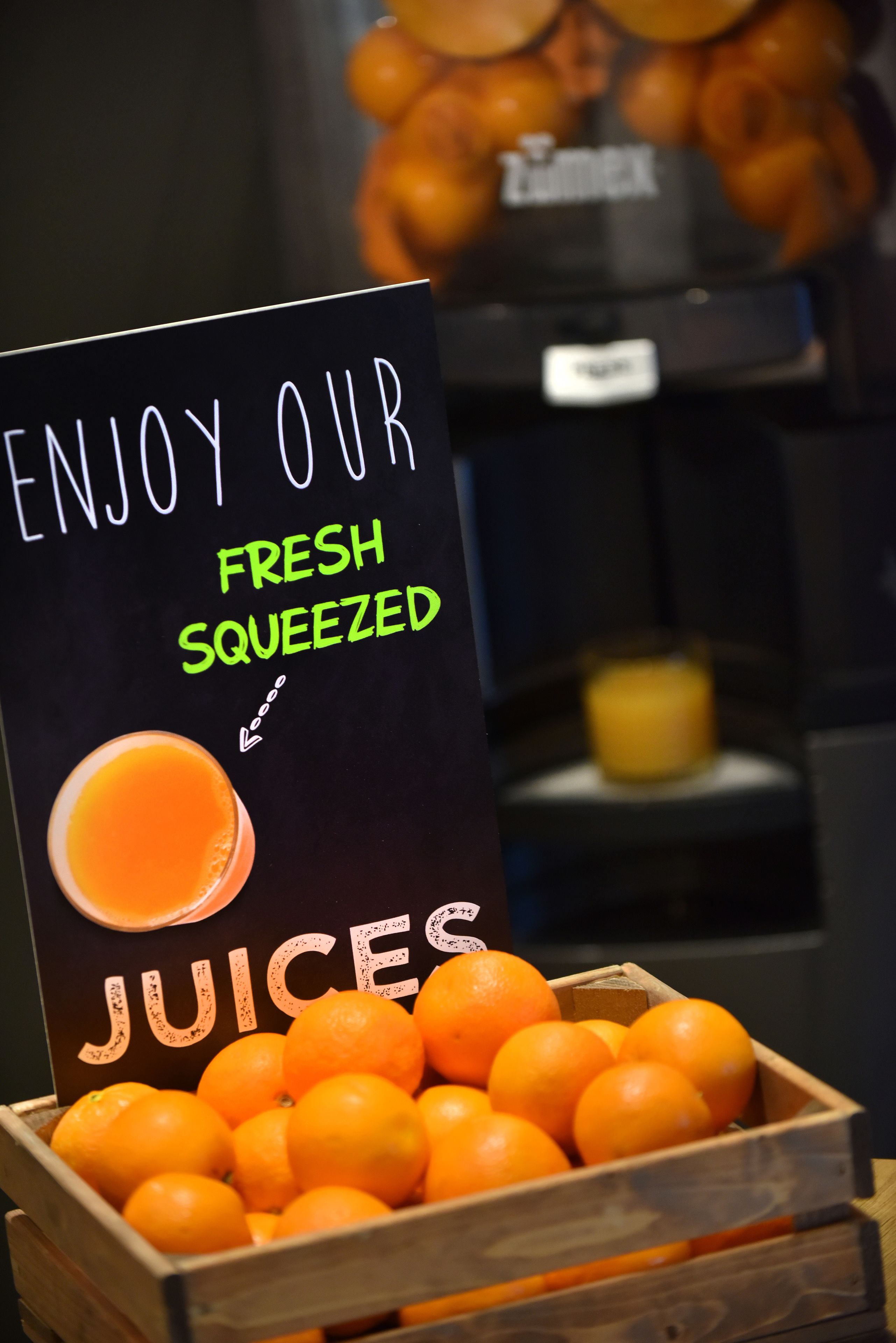 Une enseigne publicitaire pour des jus de fruits fraîchement pressés, avec une caisse en bois remplie d’oranges au premier plan.