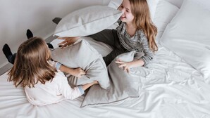 Una cama con ropa de cama y almohadas blancas, una persona sentada en la cama y otra persona acostada.