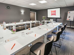 Un salón de conferencias con mesas blancas, sillas, una pantalla de proyector y un reloj en la pared.