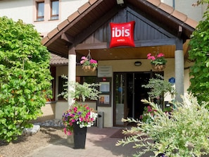A hotel entrance with a sign reading 'ibis HOTELS,' a covered entrance, and potted plants with flowers.