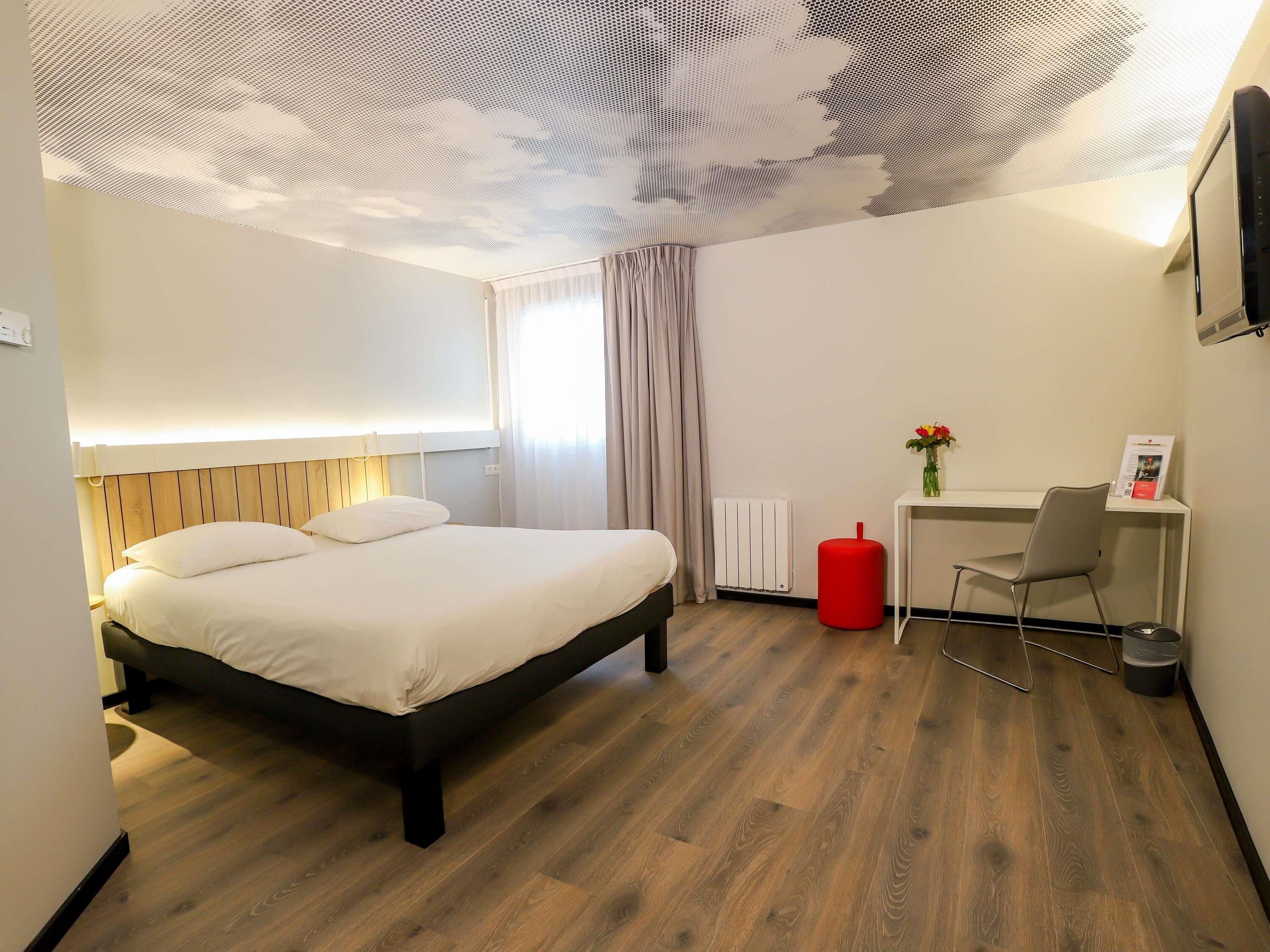 Una habitación de hotel moderna con piso de madera, una cama con sábanas blancas, un escritorio con una silla, un reposapiés rojo y un radiador.