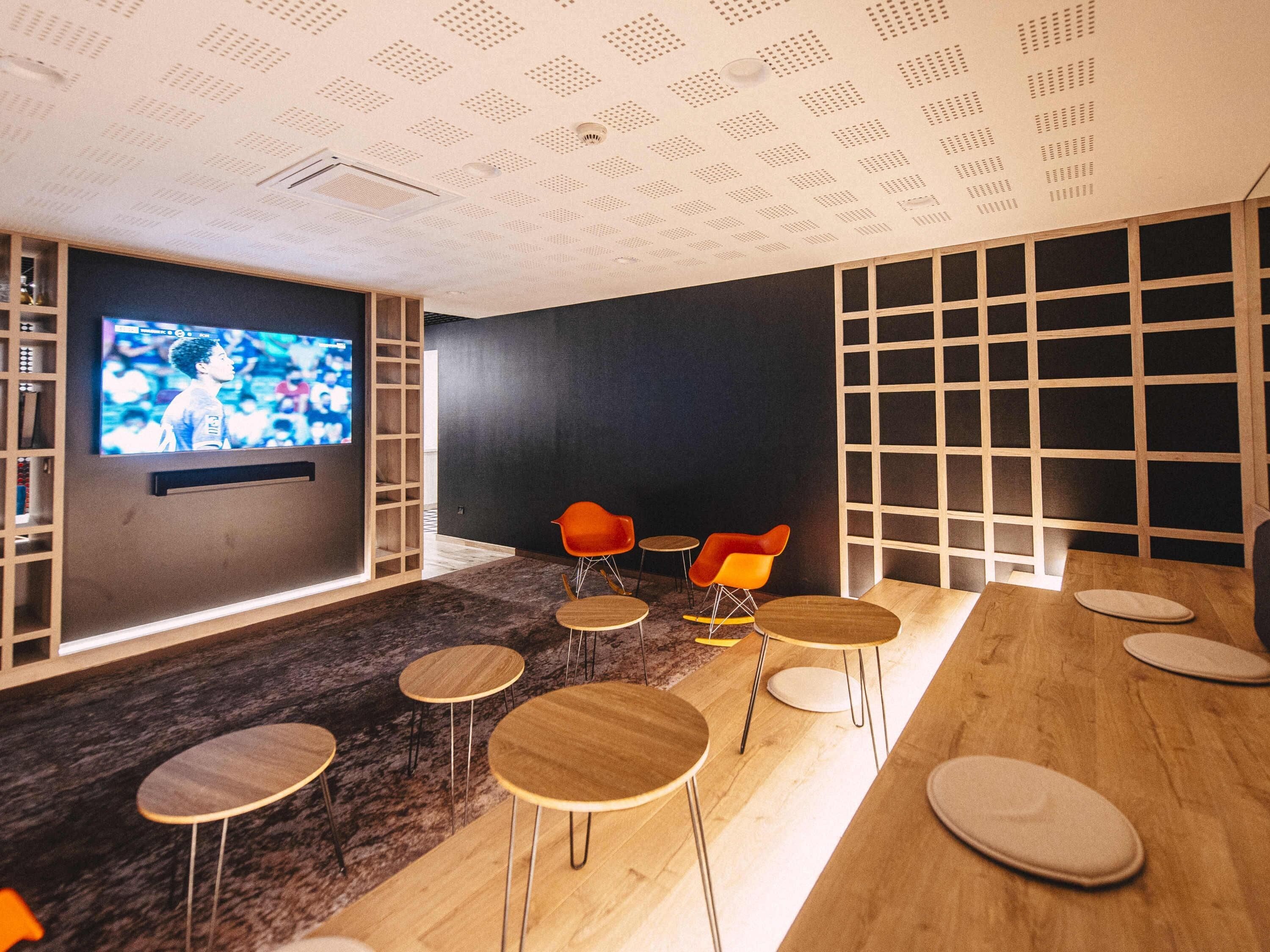 A modern lounge with a TV, wooden tables, and orange chairs.