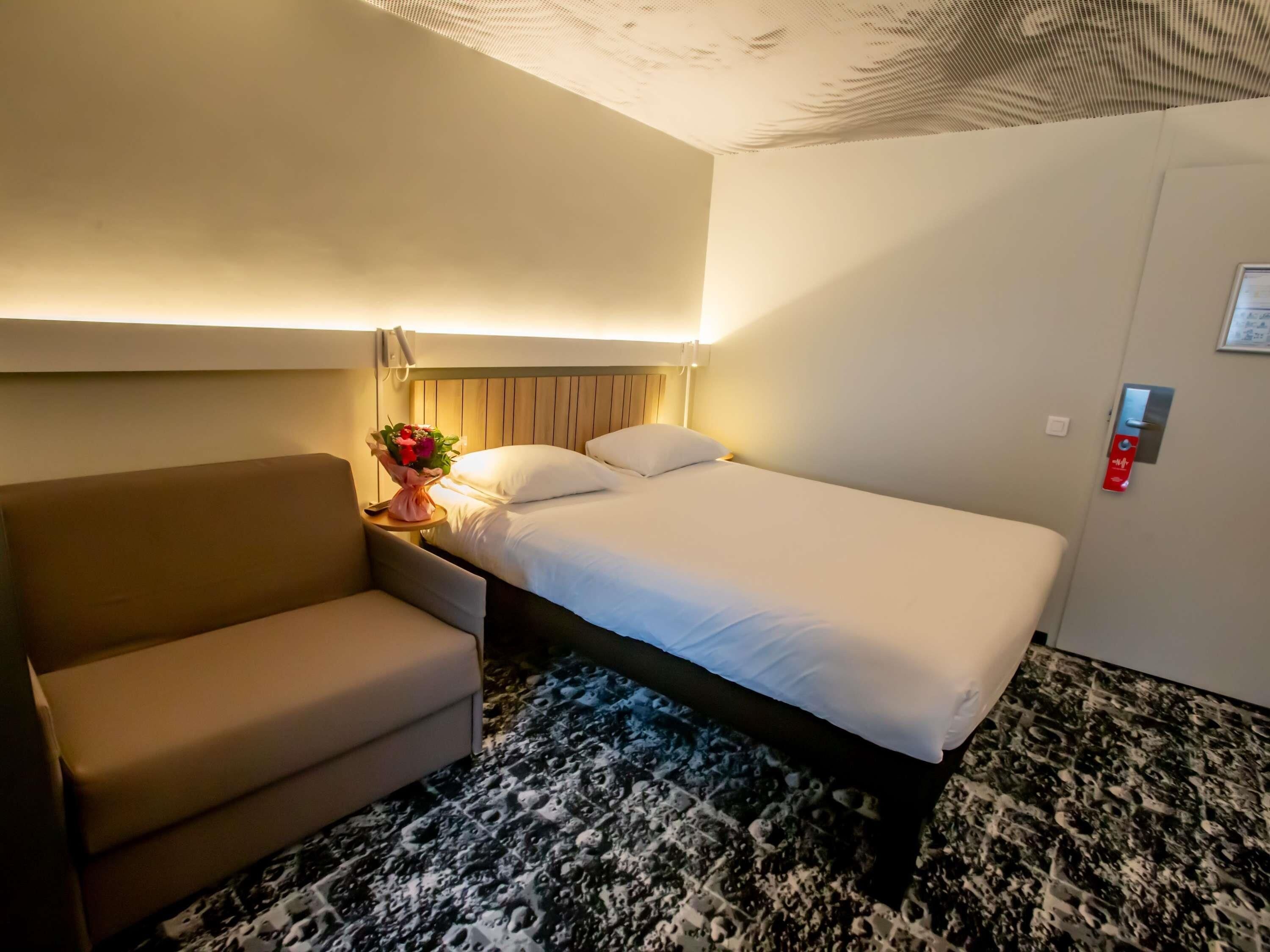 Ein Hotelzimmer mit Bett, Stuhl, Nachttisch mit Blumenvase, Wandtelefon und einer Tür mit rotem Notrufknopf.