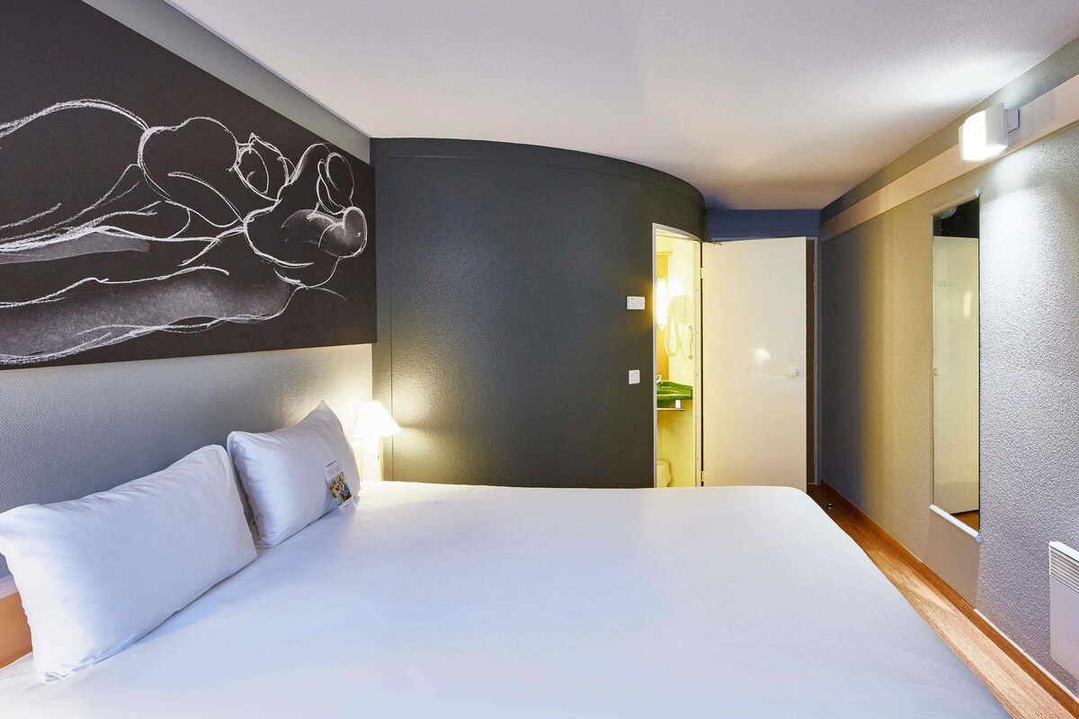 A hotel room with a large bed, a wall-mounted artwork, a bedside lamp, and a door leading to a bathroom.