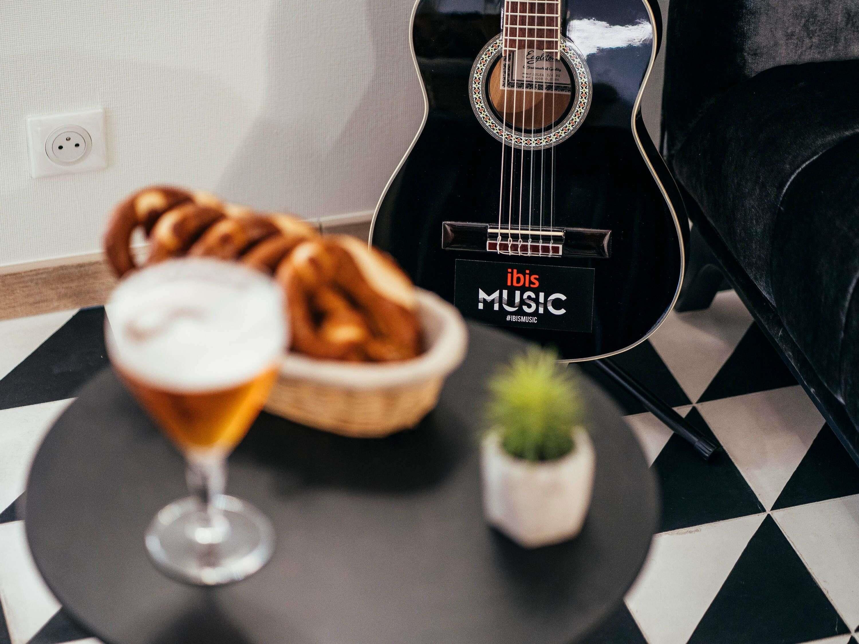 Una guitarra negra con la marca 'ibis MUSIC', un vaso con una bebida, una cesta de pan y una planta pequeña sobre una mesa a cuadros.