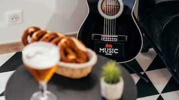 Una guitarra negra con la marca 'ibis MUSIC', un vaso con una bebida, una cesta de pan y una planta pequeña sobre una mesa a cuadros.