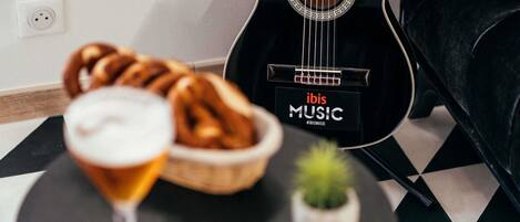 A black guitar with 'ibis MUSIC' branding, a glass with a beverage, a basket of bread, and a small plant on a checkered table.