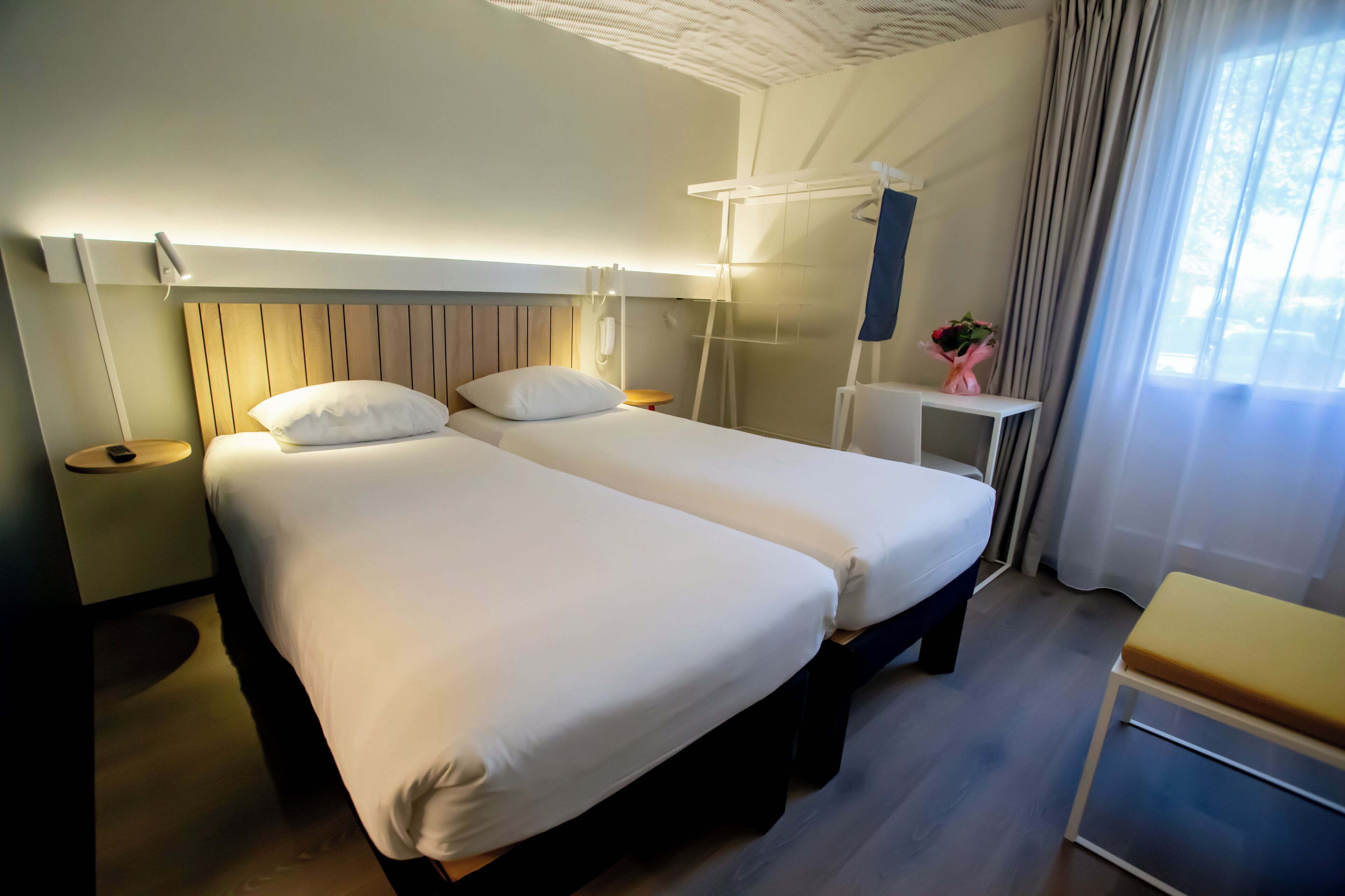 A hotel room with two beds, a bedside table, a small desk, a chair, a lamp, and a window with curtains.