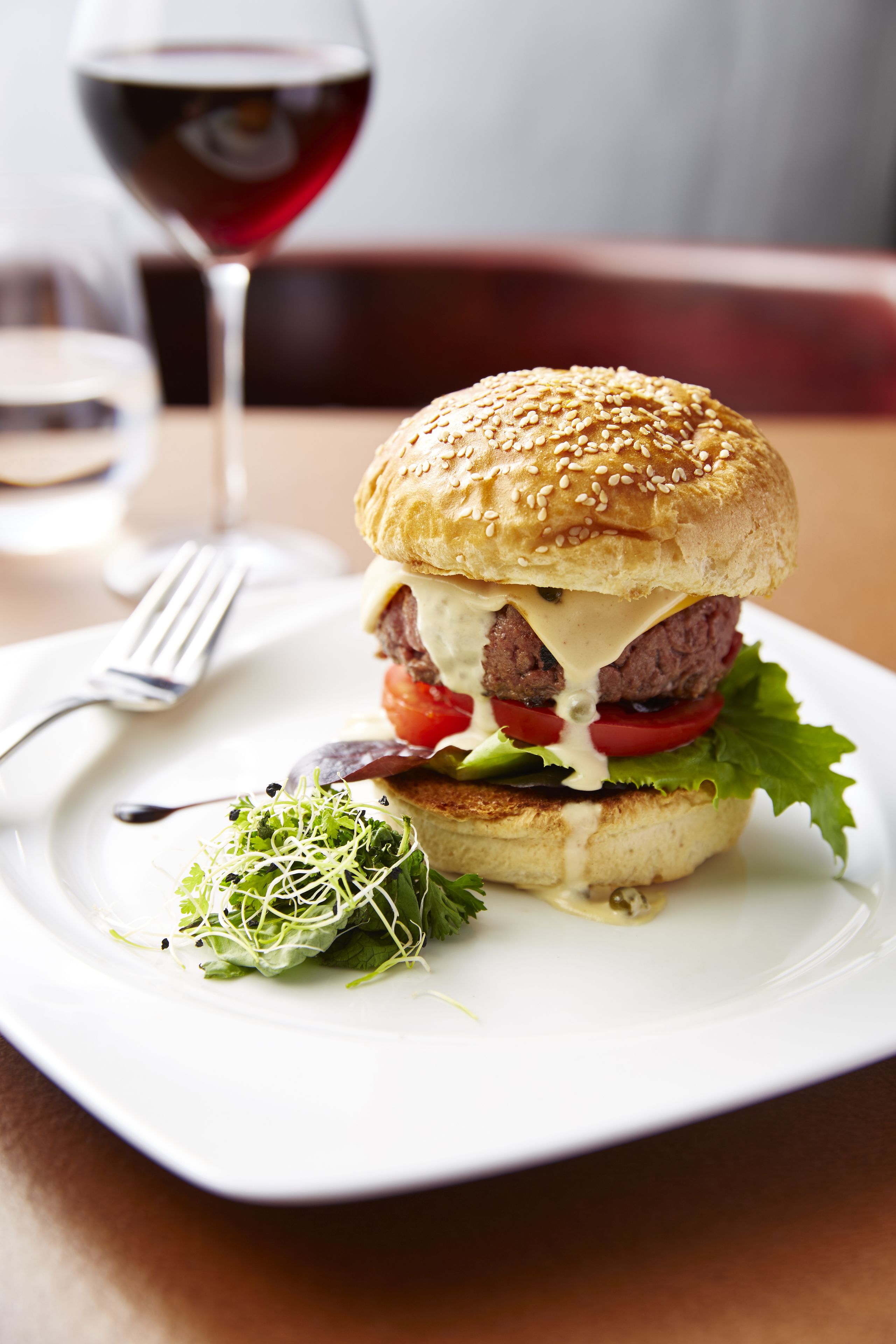 A burger with lettuce, tomato, and cheese, served on a white plate with a fork, and a glass of red wine in the background.