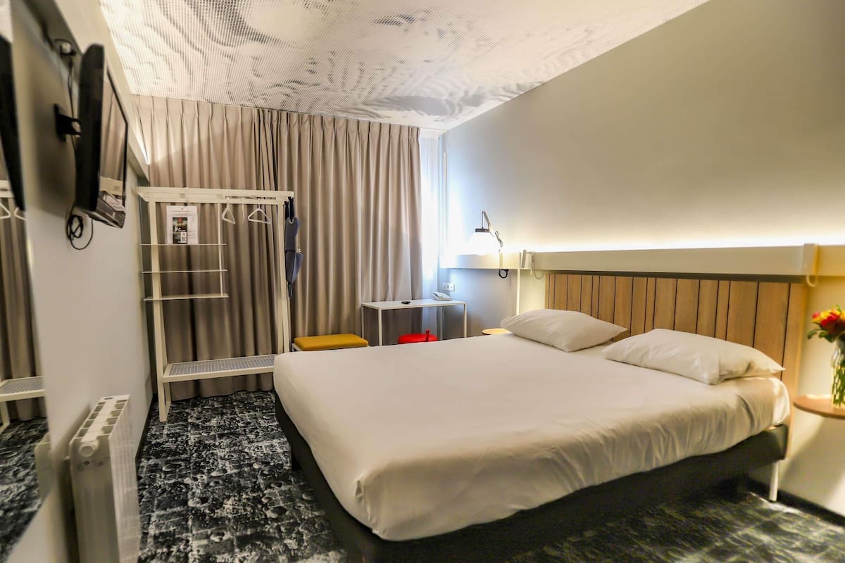 A hotel room with a bed, a desk, a chair, a wardrobe, and a radiator.