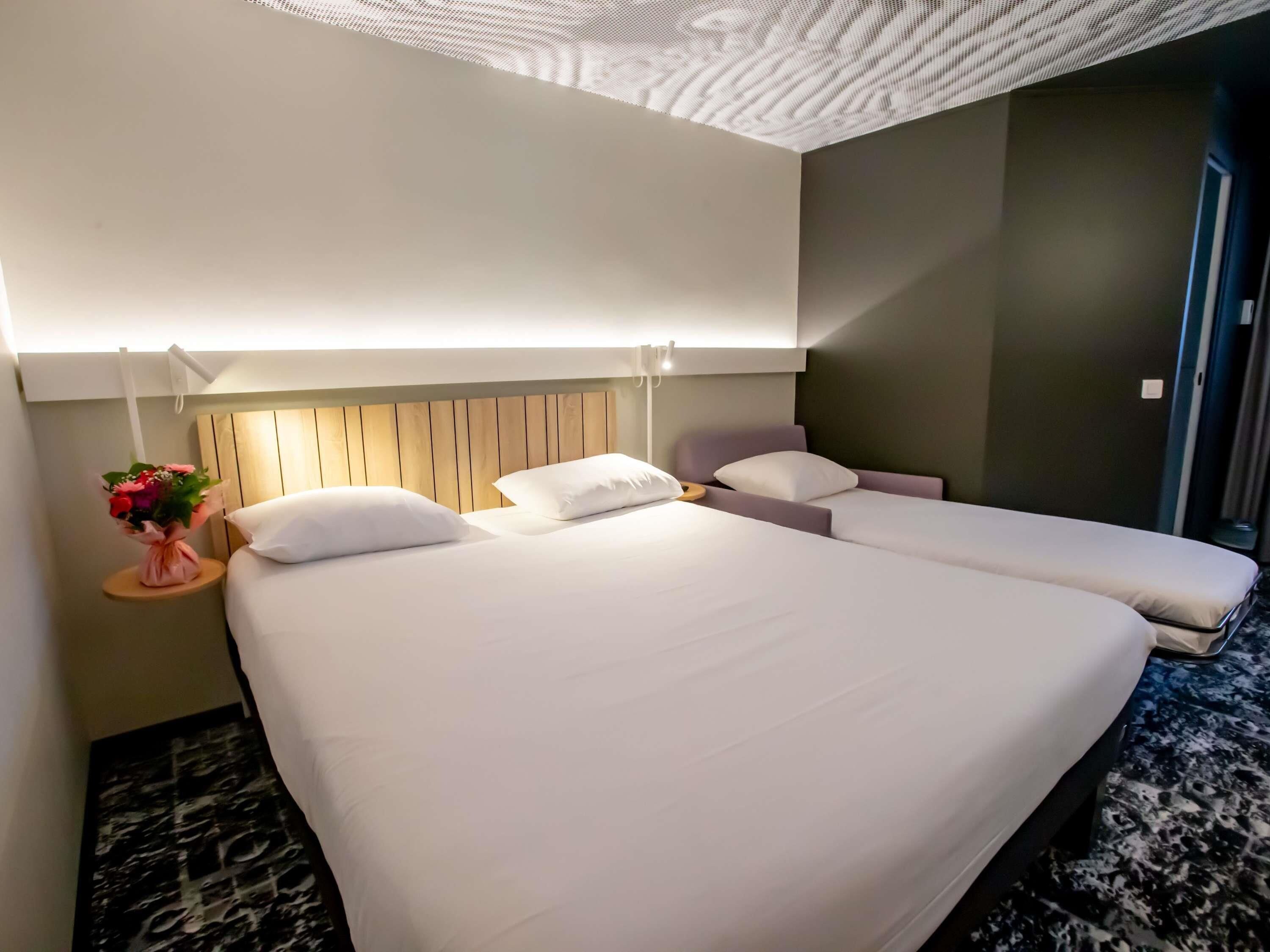 Habitación de hotel con una cama grande, una cama más pequeña, una mesita de noche con un florero y un diseño moderno en el techo.