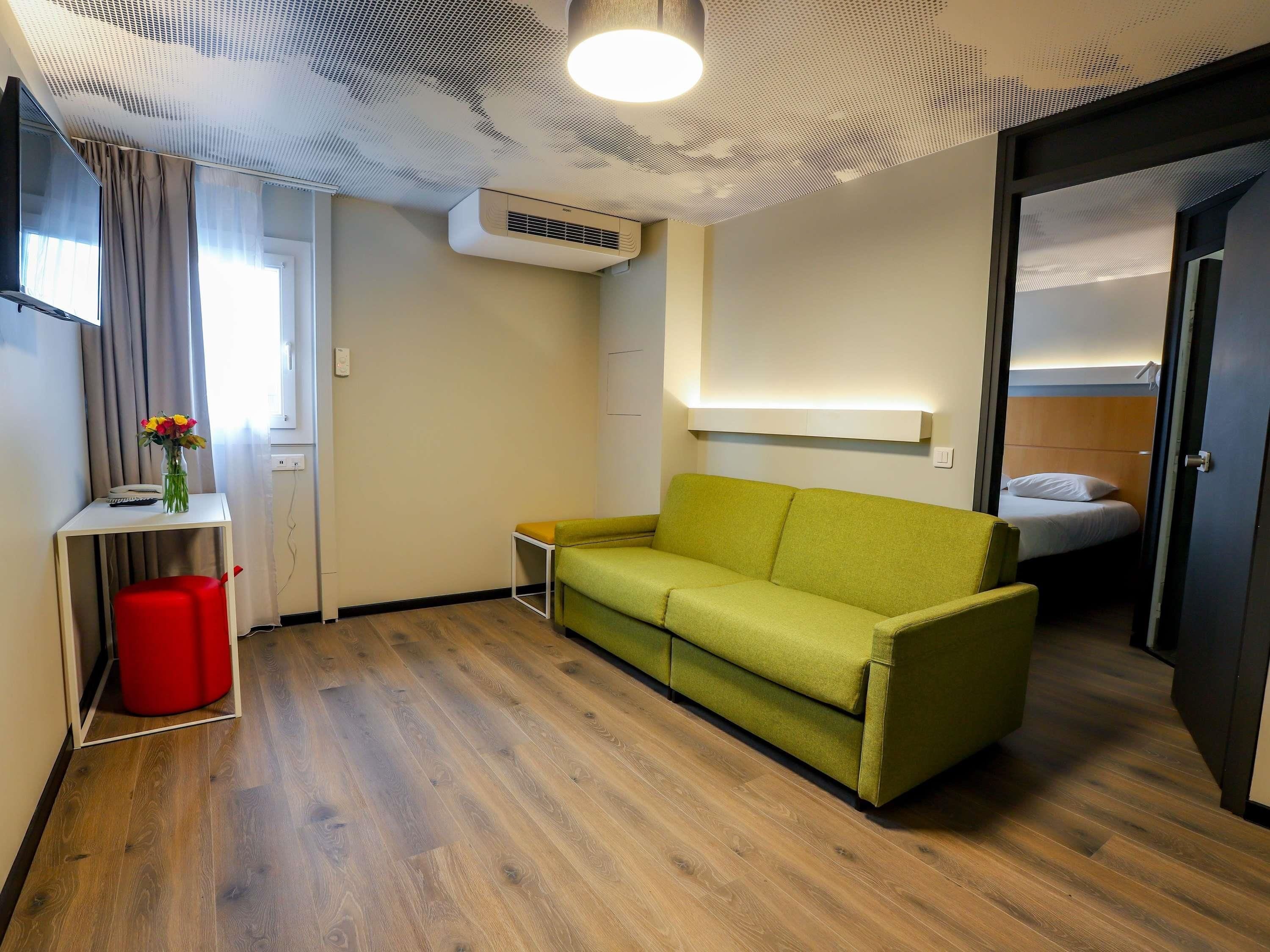 Une chambre d’hôtel moderne avec un canapé vert, un plancher en bois, une petite table avec un vase, une poubelle rouge et un lit avec une tête de lit en bois.