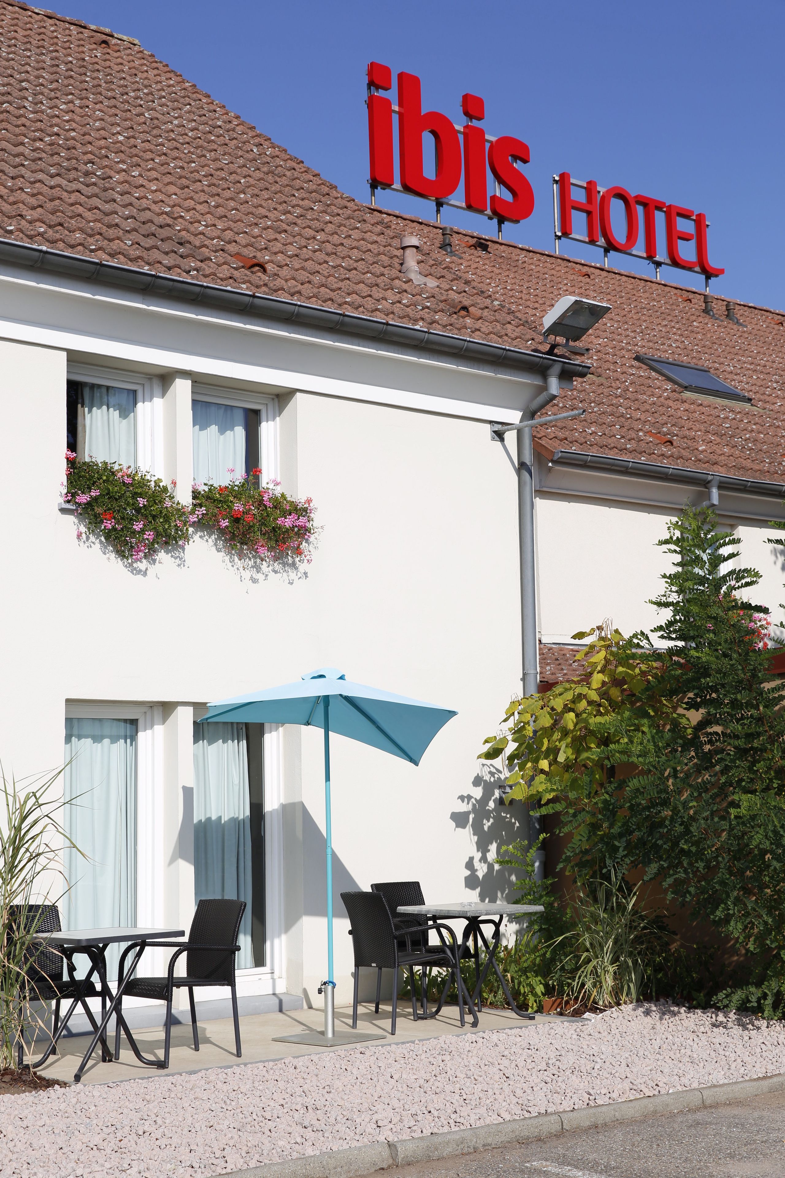 A hotel named "ibis HOTEL" with a tiled roof, outdoor seating with a blue umbrella, and flower boxes on the windows.